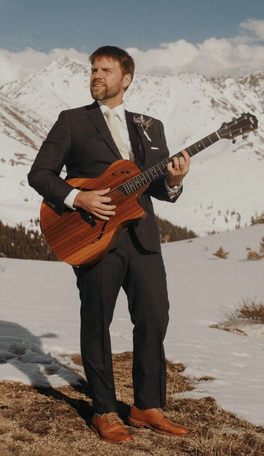 ben atop a snowy mountain in a dark suit holding a guitar